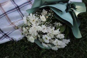 A white flower bouquet wrapped in green paper on a plaid picnic blanket outdoors.