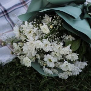 A white flower bouquet wrapped in green paper on a plaid picnic blanket outdoors.